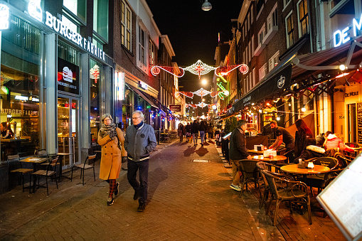 In Amsterdam, Netherlands a senior couple walks down a pedestrain street lined with people sitting outside on tables and chairs on a winter night.