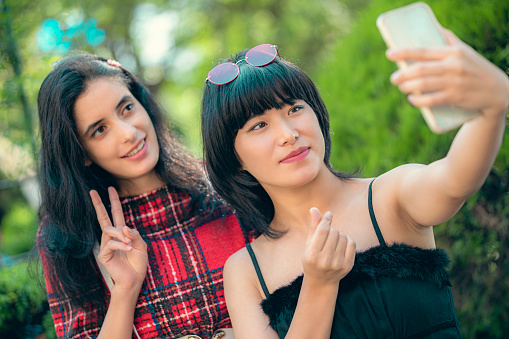Two Asian/Indian gorgeous multiracial young women capture selfies while enjoying their time in the park. These female friends wear fashionable dresses and make hand gestures while taking a selfie in the summer daytime.