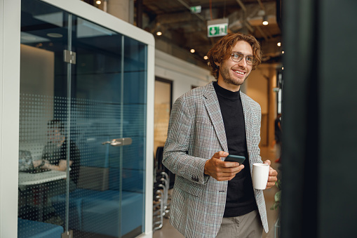 Handsome male manager use mobile phone and drink coffee while standing on office background