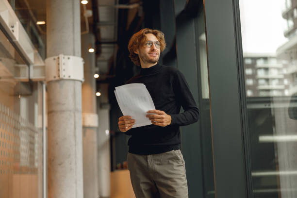 istock Smiling male entrepreneur with documents standing on modern office background and looks away 1717271102