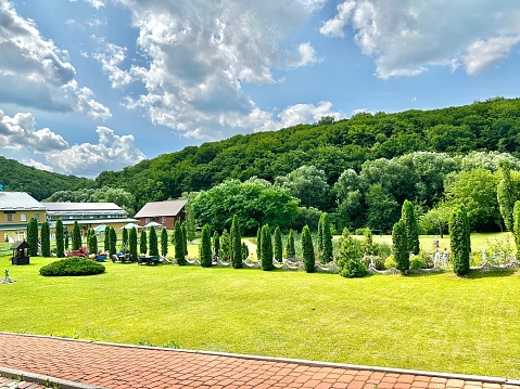 View of a park with green lawn and ornamental trees in a resort location in the middle of hills with trees on a sunny summer day and blue sky with white clouds.