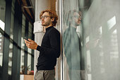 Handsome male freelancer holding phone while standing in office during break time and looks away