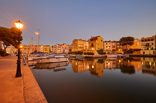 Panoramic view of colorful houses and moored yachts in the evening in Port Saplaya. Light from street lamps, buildings and boats reflects on the smooth surface of the water. Valencia's Little Venice.