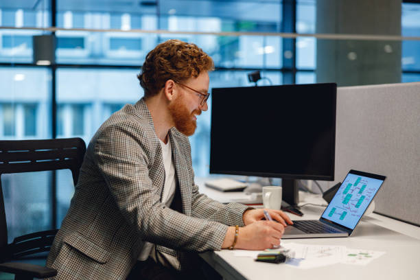 istock Bearded businessman working on laptop and making notes while sitting desk on office background 1717318015