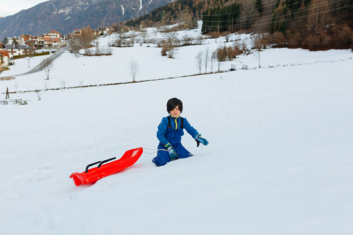 child girl with short dark hair in blue winter clothes going up the snowy hill pulling up the red sled