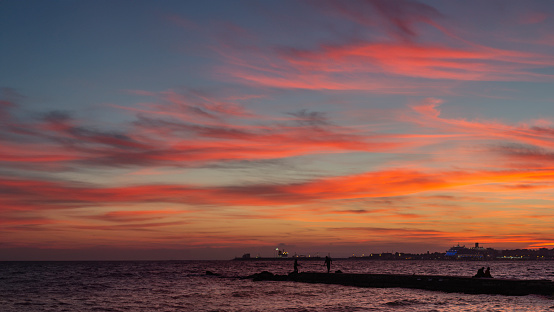 In the Bay of Palma, a sunset casts reddish hues. The sea meets a jetty with silhouetted fishermen. Far off, a cruise ship's lights dot the horizon.