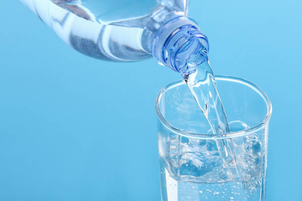 istock Pouring water from bottle into glass on light blue background, closeup 1717380849