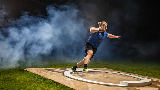 Track and field male athlete throwing shot put while training at sports court at night
