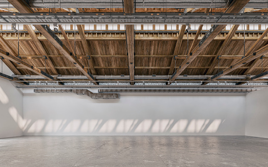 Side view of an empty large, long gallery/showroom interior with white empty walls for events or artwork exposure, and a ceiling industrial construction with hardwood beams, rooftop windows, ventilation pipes, and reflectors (lighting equipment)v on the dark gray cement tile floor. 3D rendered image.