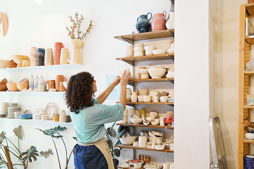 Side view of anonymous female artisan with curly hair in apron looking up while standing near shelves with ceramic products and arranging cups on wooden plank in daylight