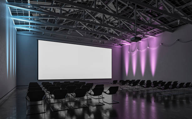 istock Side view of an empty darkened large gallery/showroom interior with empty seats (no people), and a wide projection screen in the middle for events under a ceiling industrial construction, illuminated by multi-colored side wall reflectors 1717439364