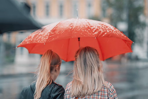 Two blonde girls under red umbrella, rear view, modern city in rain