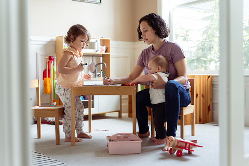 An Eurasian woman sits at a child size table at home in the playroom and does an art project with her three year old daughter while holding her 8 month old son. The woman is applying Montessori education techniques while homeschooling her preschool age daughter.