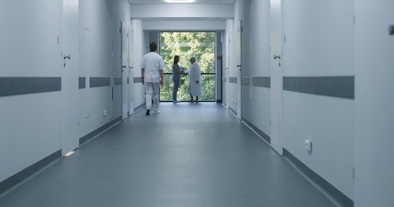 Clinic corridor: Doctors and professional medics walk. Nurse with digital tablet comes to elderly woman standing near window. Medical staff and patients in modern hospital or medical center hallway.