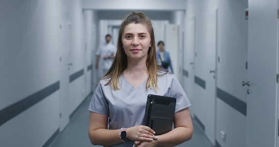 Female doctor stands in modern clinic corridor. Professional medic holds digital tablet, smiles and looks at camera. Medical staff and patients in background in hospital or medical center hallway.