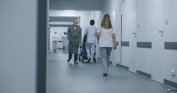 Mature doctor stands in clinic corridor with nurse and patient. Professional medic talks with female colleague and elderly woman, uses digital tablet. Medical staff and patients in hospital hallway.