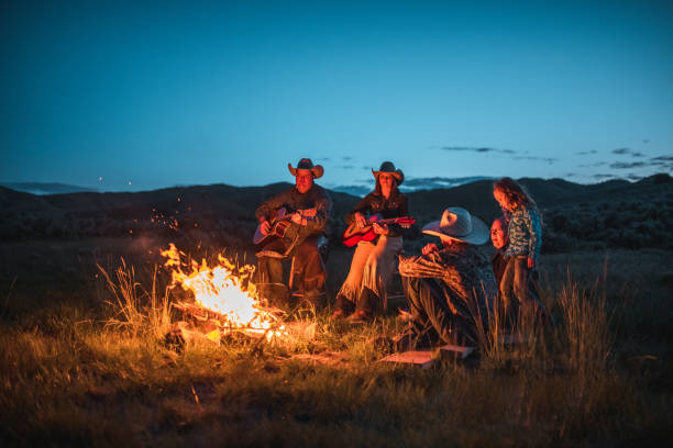 Western american cowboys and cowgirls gathering together while listening to the melodies coming from the guitarist. They are seated next to a campfire during the blue hour.