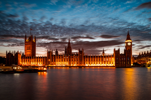Waterfront view of the illuminated Houses of Parliament and Big Ben at dusk with a dramatic sky.