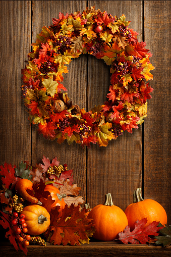 A Thanksgiving wreath hangs on a barn wood wall as pumpkins, gourds, and autumn leaves are gathered below.