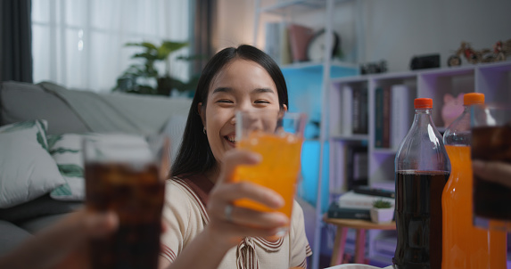 Selective focus Asian young woman and her friend clink a glass of beverage while party in living room at home, They are happiness during celebration of friendship