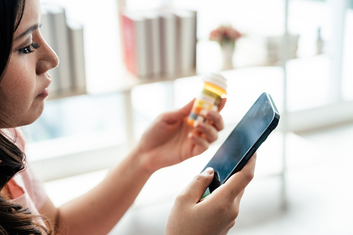 Young woman holding pills bottle and searching medications dosage instruction online on smartphone in living room