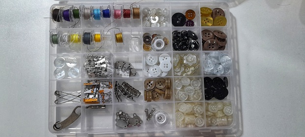 A plastic sewing box set with all essential sewing  items i.e a variety of buttons, sewing thread, hook for sewing process. A closeup photo of a professional sewing kit with sewing items on white back ground.