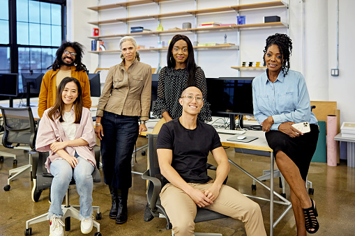 Diverse group in 20s, 30s, and 50s, casually dressed, sitting and standing at flexible workstation in modern loft office, smiling at camera.