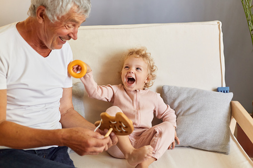 Extremely happy baby granddaughter posing with gray haired mature man wearing white T-shirt sitting on sofa in living room at home playing with toys having fun together.
