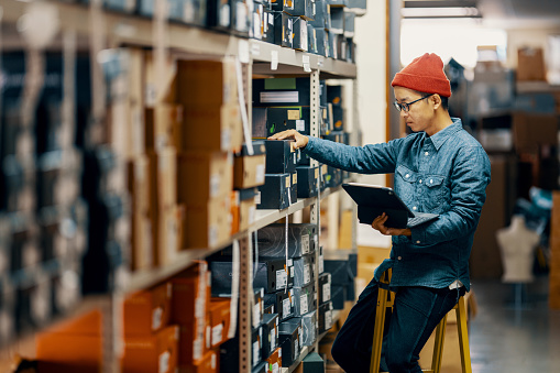 Mid adult male retail shop staff checking or looking for inventory in a storage room by using a digital tablet