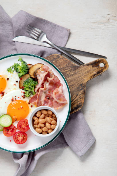 plato de desayuno tradicional englis con tiras de tocino, huevos soleados, verduras y pastel sobre fondo claro - englis fotografías e imágenes de stock