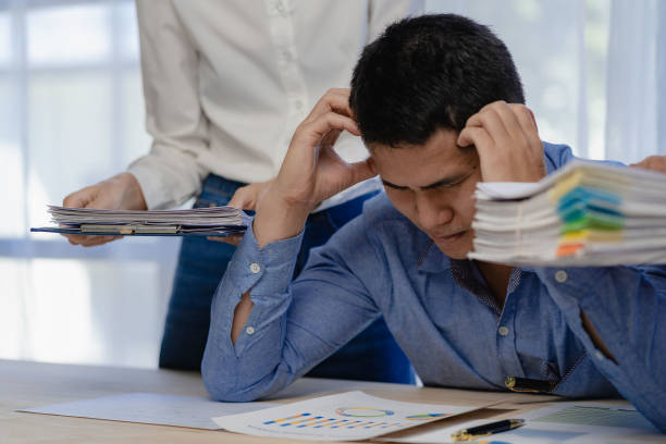 istock Stressed businessman sitting at workplace and touching his headYoung asian exhausted businesssman with messy desk and stack of papers, working busy, overwork. 1718141846