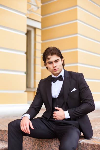 istock Confident businessman guy in tuxedo sitting outdoors, strong looking at camera. Serious handsome young man in tux posing at urban street outside, luxury image James Bond style. Copy ad text space 1718145558