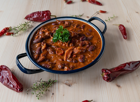 Delicious kidney bean stew with smoked pork meat and vegetables, tex mex style. Served hot and ready to eat in a rustic bowl isolated on light wooden background with dried chili peppers.