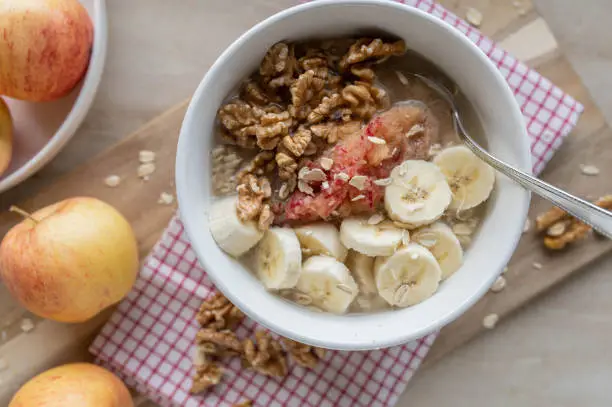 Natural and vegan  breakfast oatmeal with soaked oats, walnuts, grated apples, bananas and honey. Served ready to eat in a bowl with spoon on light pine wood background from above. Lactose free