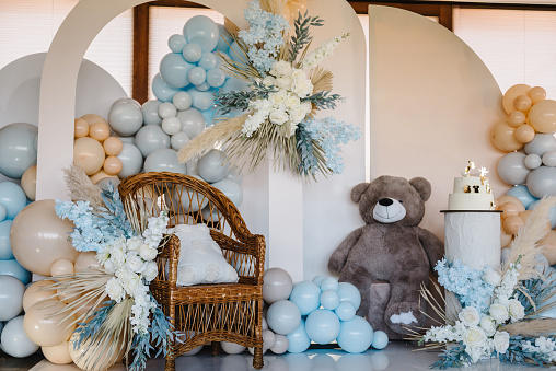 Arch with bear, wicker wooden chair. Trendy cake for celebration baptism. Delicious reception at birthday party. Photo wall decoration blue, brown, grey balloons, autumn decor with dry leaves, flowers