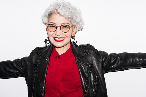 A gray-haired senior Japanese woman is smiling at the camera.She wears sunglasses, a leather jacket and large earrings.Studio shot on white background.