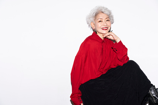 A gray-haired senior Japanese woman is smiling at the camera.She is wearing red clothes and big earrings.Studio shot on white background.