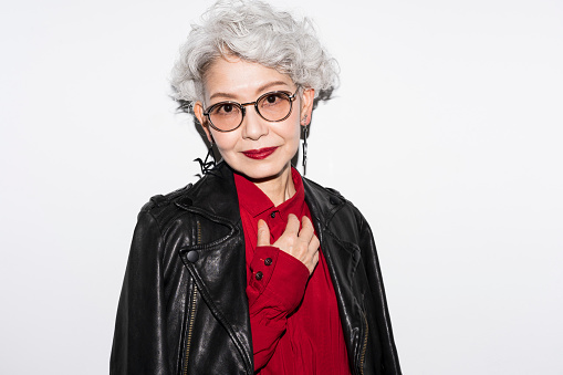 A gray-haired senior Japanese woman is smiling at the camera.She wears sunglasses, a leather jacket and large earrings.Studio shot on white background.