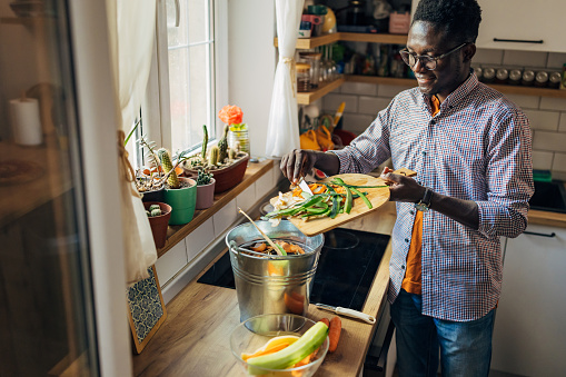 A black man learning how to cook in a domestic kitchen with fruits and vegetables. He is preparing a healthy vegan or vegetarian meal with organic ingredients and throw organic waste in compost bin