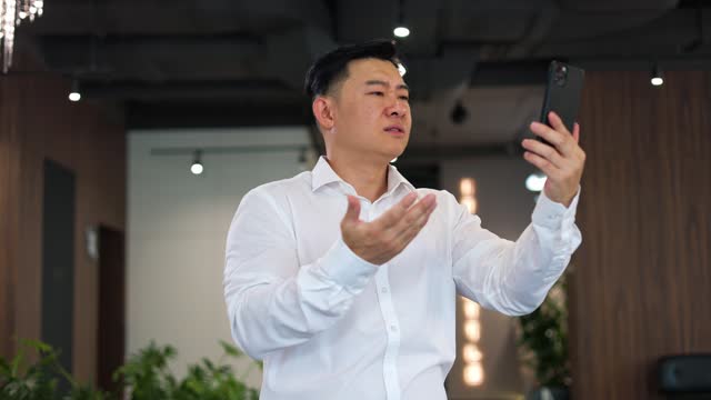 Anxious businessman facing low connectivity issues while staying in corporate office. Annoyed asian gentleman in formal attire raising smartphone up and moving it in order to catch wi-fi signal.