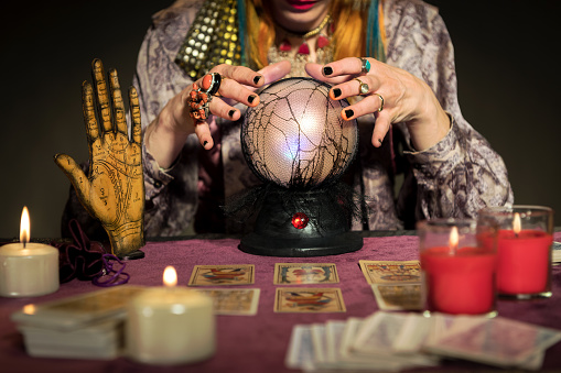 Crop anonymous fortune teller female looking down while sitting at table with hovering fingers on crystal ball burning candles blurred cards and palmistry reading picture