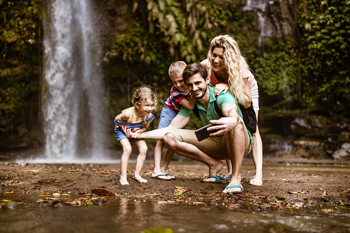 Young happy family taking a selfie with cell phone by the waterfall in rainforest.