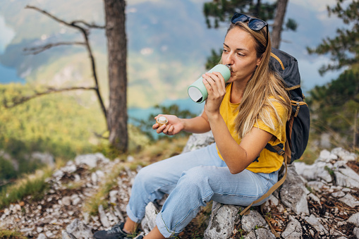Female tourist sitting on top of mountain above canyon resting after climbing drinking water from reusable bottle