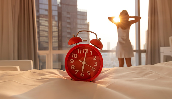 Red alarm clock with Fresh of young woman open a curtains and stretching in bed after waking up, sunlight in morning