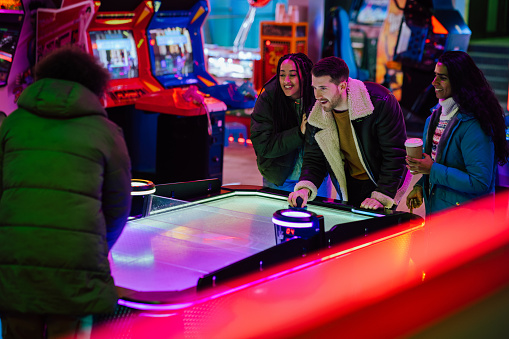 A group of friends on a double date, spending the day at an amusement arcade together during a day in winter in Whitby, North East England. They are playing air hockey together and one man is holding a handheld disc on the table, ready to receive the plastic puck from his opponent while two of the women stand next to him and watch.\n\nVideos are also available for this scenario.