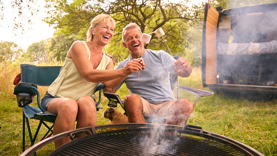 Senior Couple Camping In Countryside With RV Toasting Marshmallows Outdoors On Fire