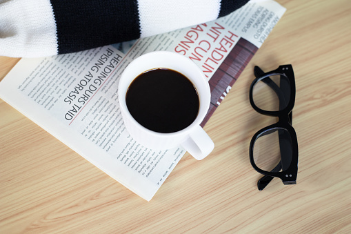 A cup of black coffee with newspaper, scarf and eyeglasses on wooden table
