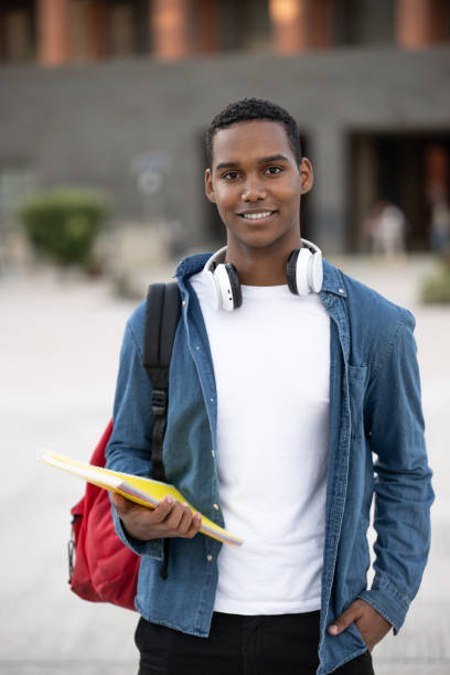 istock Handsome student wearing a bacpack and holding a notebook looking at camera.Relaxed and happy man standing outside with headphones ready for college. 1718413021