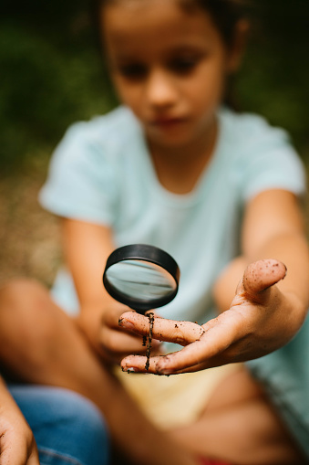 Girl sitting in nature and looking worms with magnifying glass for the biology class project