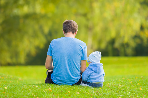 Young adult father and baby boy sitting together on green grass at park. Spending time together in beautiful autumn day. Back view. Lovely emotional moment. Peaceful atmosphere in nature. Closeup.